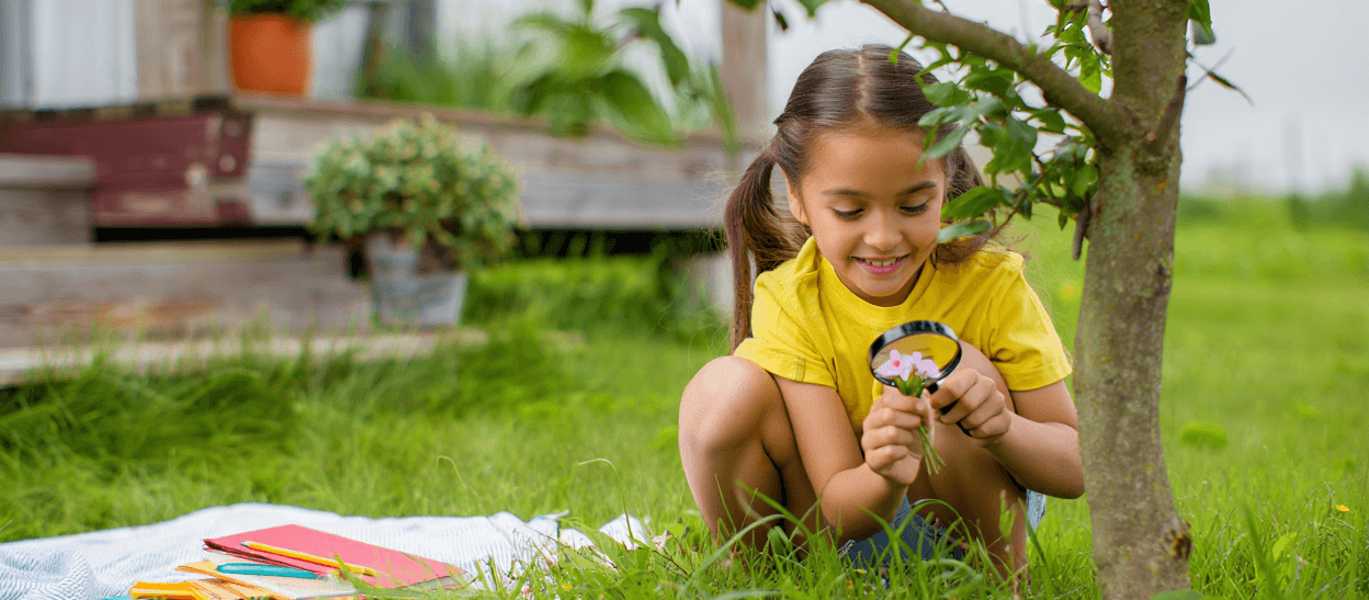 Child with pigtails examining pink flowers with a magnifying glass in grass.