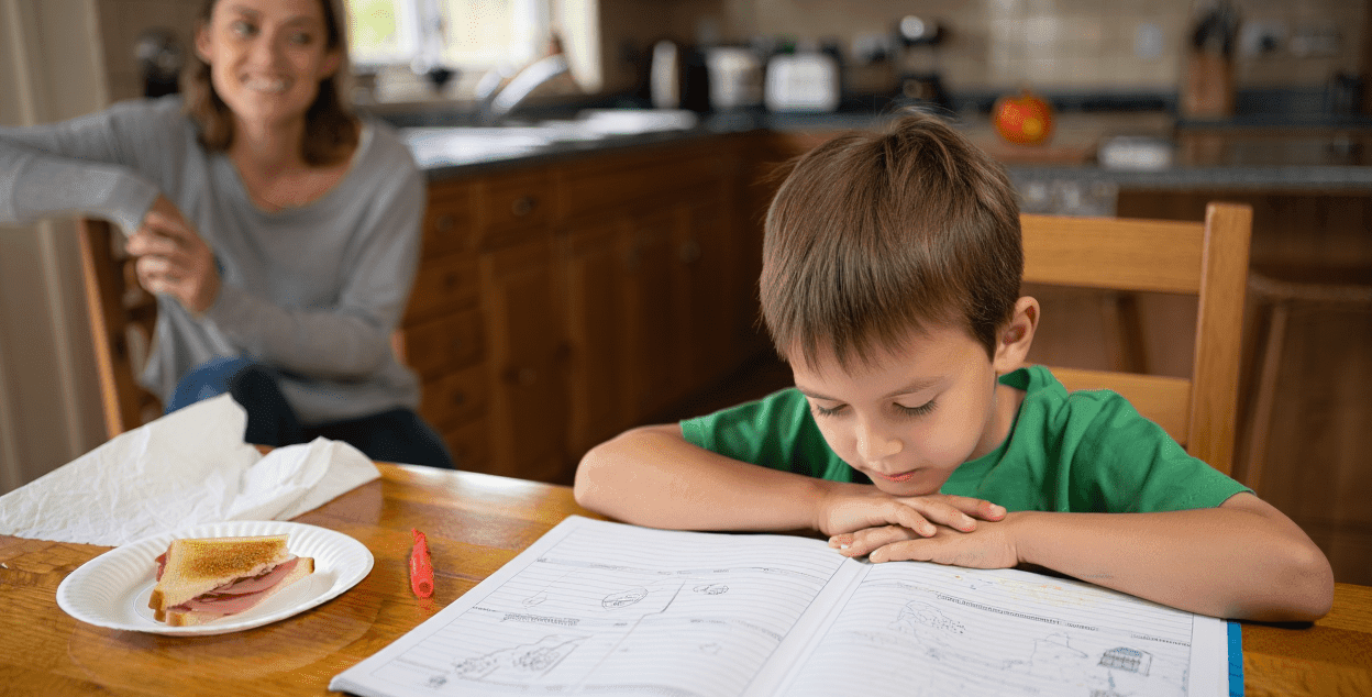 Child studying at a table with an adult smiling in the background.